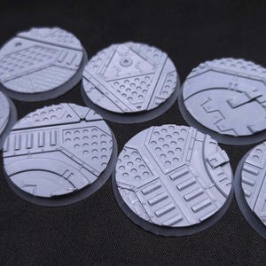 May include: A collection of eight gray, circular bases with intricate, industrial-themed surface designs. Each base is approximately 40mm in diameter, featuring patterns of panels, rivets, and geometric shapes. The bases are designed for miniature gaming.