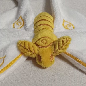 May include: A white plush hooded garment with gold embroidered accents and a yellow bee-like appliqué. The bee has detailed wings and a central eye design. The hood has gold embroidered teardrop shapes.