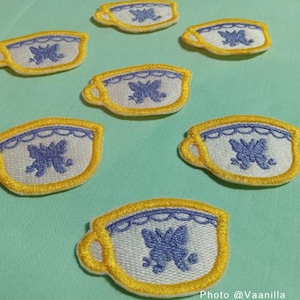 May include: Seven embroidered iron-on patches of teacups with a yellow border and a purple butterfly design. The teacups are white with a blue trim.