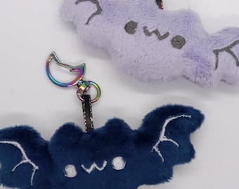May include: Two plush bat keychains, one purple and one blue, with embroidered eyes and wings. Both have a rainbow-colored metal clasp.