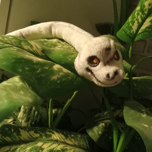 May include: A white plush snake with black eyes and a black mouth, curled around a green leafy plant.