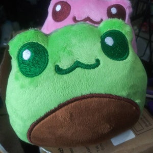 Peut inclure: Une grenouille en peluche verte avec du feutre brun sur le dessous et des yeux verts brodés. La grenouille est posée sur une grenouille en peluche rose avec des yeux bruns brodés.