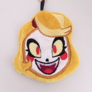 May include: Yellow plush keychain featuring a cartoon character with red eyes, a wide smile, and fangs. The character has blonde hair and is wearing a red and white outfit.