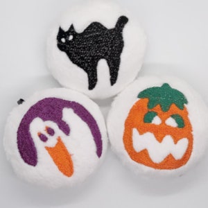 May include: Three white plush ornaments with embroidered Halloween designs. One ornament features a black cat, another a purple ghost, and the third a smiling orange pumpkin.