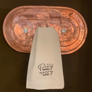 May include: A white kitchen towel with the text "Pray More Worry Less" in black cursive lettering. The towel is hanging on a copper towel rack with two crystal knobs.
