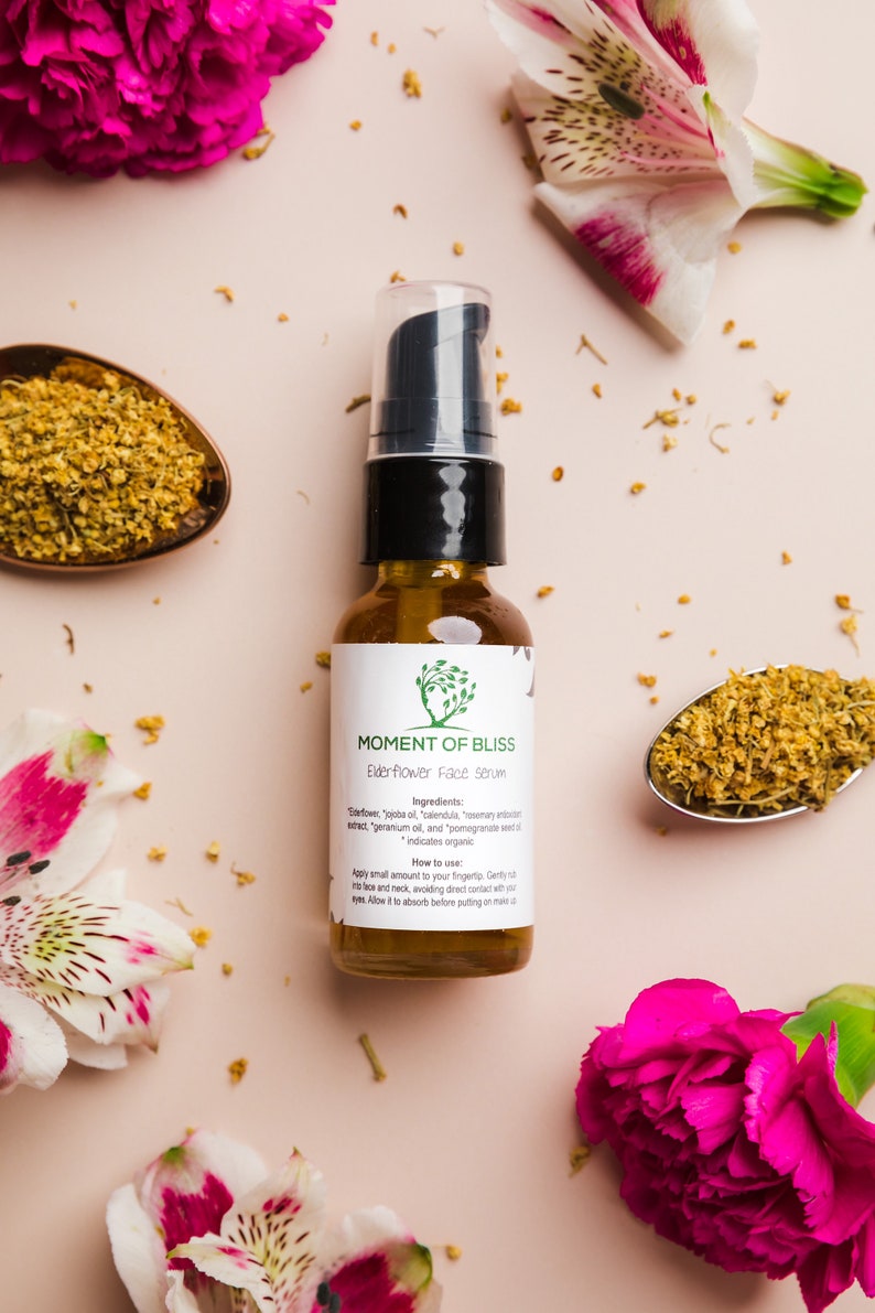 May include: A glass bottle of elderflower face serum with a white label that says "Moment of Bliss" and lists the ingredients and how to use the serum. The bottle is surrounded by pink and white flowers and a spoon with dried flowers.