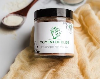 May include: A glass jar with a black lid containing a brown powder. The label reads "Moment of Bliss Dry Shampoo for dark hair". The jar is sitting on a beige fabric.