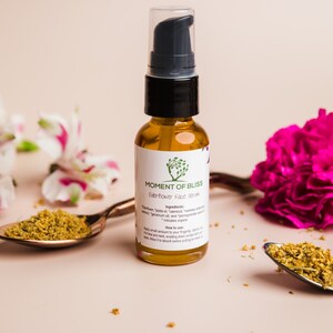 May include: A glass bottle of Moment of Bliss Elderflower Face Serum with a black pump top. The bottle is sitting on a light pink surface with dried flowers and a copper spoon.