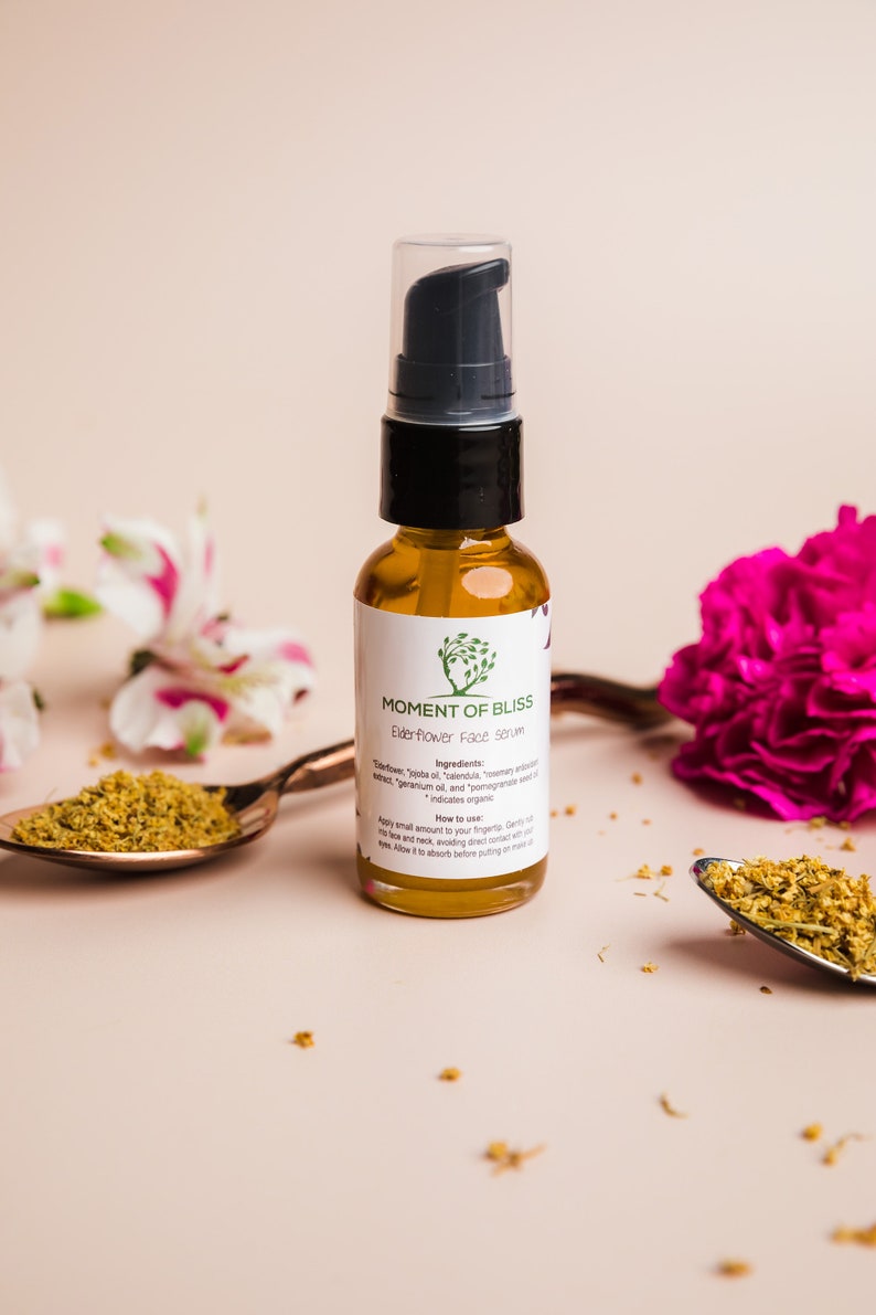 May include: A glass bottle of Moment of Bliss Elderflower Face Serum with a black pump top. The bottle is sitting on a light pink surface with dried flowers and a copper spoon.