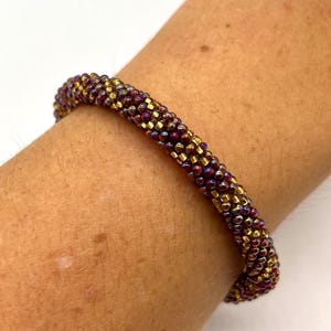 May include: A close-up of a beaded bracelet with a gradient of burgundy and gold beads. The bracelet is worn on a person's wrist.