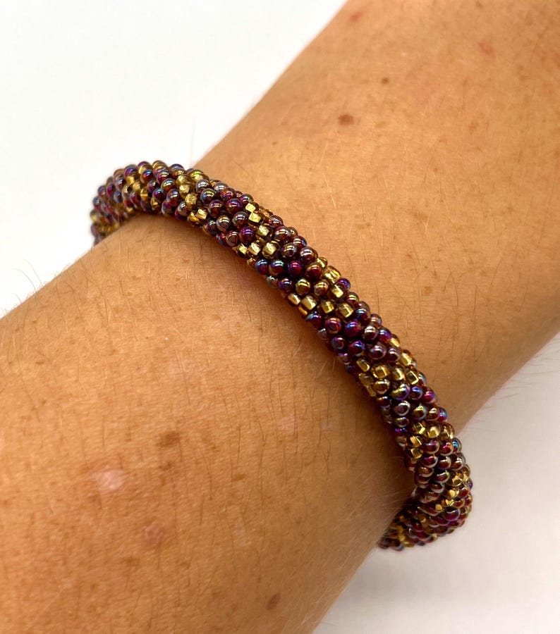 May include: A close-up of a beaded bracelet with a gradient of burgundy and gold beads. The bracelet is worn on a person's wrist.