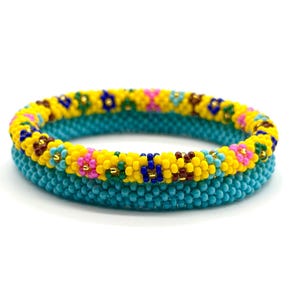 May include: Two beaded bracelets. The bottom bracelet is turquoise, and the top bracelet is yellow with a floral pattern of pink, blue, and brown beads. The bracelets are stacked on top of each other against a white background.