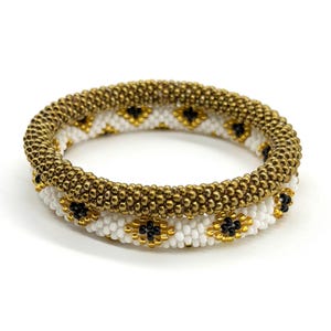 May include: Two beaded bracelets. The top bracelet is gold, and the bottom bracelet is white with gold and black accents. The bracelets are made of small beads and have a textured appearance. The bracelets are stacked on top of each other.