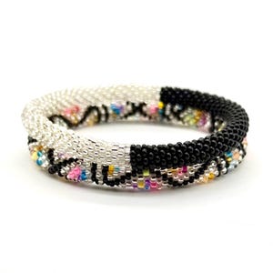Könnte beinhalten: Zwei Armbänder aus Perlen. Eines ist silber und weiß, das andere ist schwarz. Beide Armbänder haben bunte Perlen dazwischen. Die Armbänder bestehen aus kleinen, runden Perlen und sind miteinander verdreht.