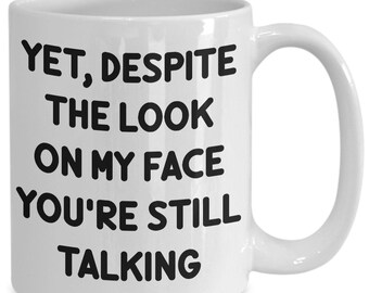 May include: White ceramic coffee mug with black text that reads "Yet, despite the look on my face you're still talking."