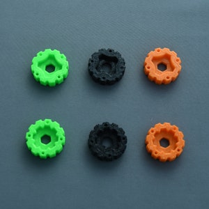 May include: Six plastic gears, two green, two orange, and two black, arranged in two rows of three on a gray surface.