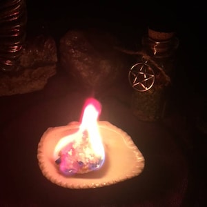 May include: A white ceramic dish with a small, white, crystalline substance burning with a bright orange and blue flame. The dish is on a dark purple surface. A small glass bottle with a cork top and a pentagram charm is in the background.