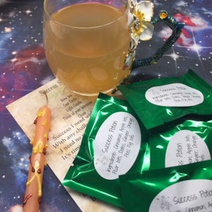 May include: Four green pouches with white labels that read "Success Potion" and list ingredients like cinnamon, apple bits, pear bits, dates, ginger, and orange. The pouches are on a table with a glass of tea, a candle, and a piece of paper with the words "Success Spell" written on it.