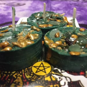 May include: Three green tealight candles with gold flecks and clear glass pieces embedded in the wax. The candles are sitting on a card with a pentagram symbol.