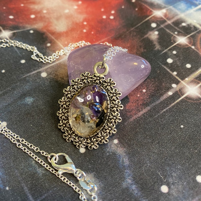 May include: A silver necklace with a pendant featuring a clear oval resin filled with amethyst, clear quartz, and gold flakes. The pendant is surrounded by a silver filigree design with small flower accents.