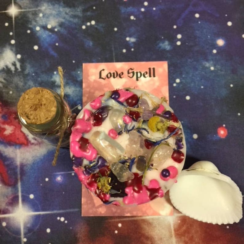 Puede incluir: Una vela rosa y blanca con cristales, hierbas y flores incrustados en la cera. La vela est&aacute; etiquetada como "Love Spell". Hay una peque&ntilde;a botella de vidrio con un tap&oacute;n de corcho y una concha blanca.