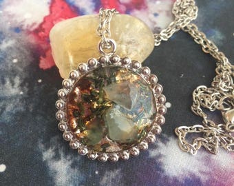 May include: A silver pendant necklace with a round, clear resin pendant. The pendant contains green and gold inclusions, and is surrounded by a beaded silver frame. The necklace chain is also silver.