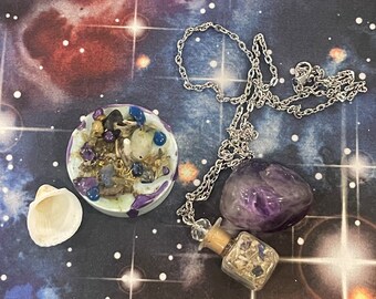 May include: A silver chain necklace with a purple amethyst crystal pendant and a small glass bottle filled with crystals. The necklace is displayed on a purple and white background with a small white seashell and a purple and white bowl filled with crystals.