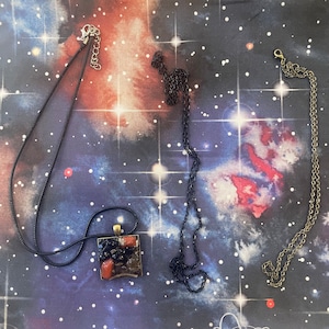 May include: Two necklaces with pendants. One necklace is black with a square pendant with red, black, and brown stones. The other necklace is gold with a simple design.