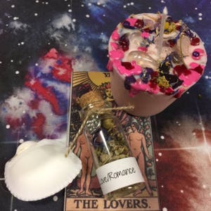 Puede incluir: Una vela rosa con flores secas y cristales, una peque&ntilde;a botella de vidrio con hierbas secas y una etiqueta que dice "Love/Romance", y una concha blanca. Los art&iacute;culos est&aacute;n dispuestos en una carta de tarot con la imagen de Los Amantes.
