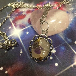 May include: A silver chain necklace with a pendant featuring a clear quartz crystal with small amethyst crystals inside. The pendant is set in a silver frame with a decorative scalloped edge. The necklace is displayed on a pink rose quartz heart.