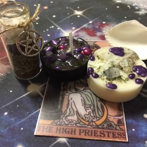 May include: Two small candles, one black with purple accents and one white with purple accents, are sitting on a tarot card that reads "The High Priestess". A small glass bottle with a pentagram charm is also in the image.