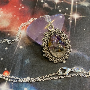 May include: A silver chain necklace with a pendant featuring a clear resin oval filled with purple and white crystals, surrounded by a silver floral design.