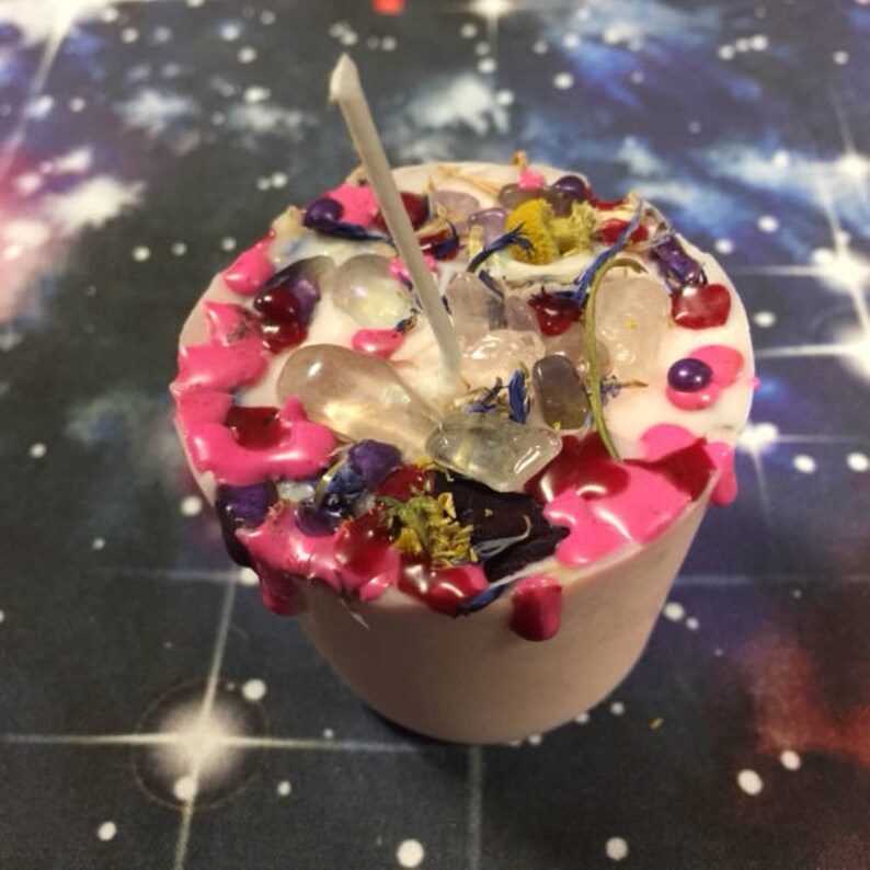 Puede incluir: Una vela rosa con una mecha blanca y cristales, flores y cuentas incrustados. La vela est&aacute; sobre un fondo oscuro con un patr&oacute;n estrellado.