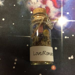 Puede incluir: Una peque&ntilde;a botella de vidrio con un tap&oacute;n de corcho llena de flores y hierbas secas. La botella tiene una etiqueta blanca con el texto "Love/Romance". La botella est&aacute; decorada con un amuleto plateado que dice "With Love".