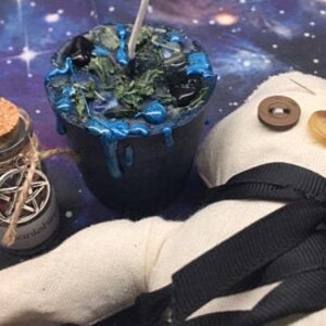 May include: A black candle with blue wax dripping down the sides. The candle is surrounded by green and blue crystals and a small bottle with a cork stopper and a pentagram symbol on the label. A white fabric doll with black ribbon and buttons is in the background.