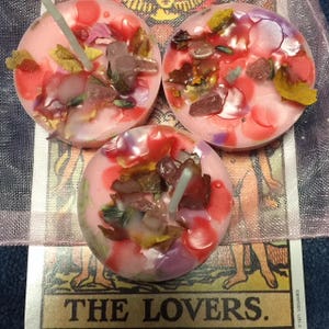 May include: Three pink round candles with embedded crystals and dried flowers. The candles are on top of a tarot card with the text "THE LOVERS."