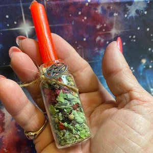 May include: A glass bottle filled with dried herbs and flowers, topped with a red candle and a silver pentagram charm. The bottle is held in a hand.