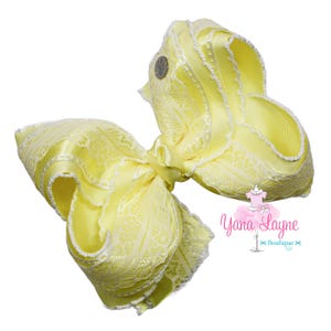 May include: A large, yellow hair bow with white lace trim and a small, silver emblem. The bow is made of layered satin ribbon and features a central knot. The text "Yana Layne Boutique" is visible.