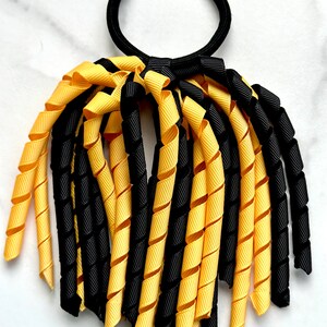 May include: Black and gold curly ribbon hair tie. The hair tie is made of a black elastic band with multiple strands of black and gold curly ribbon attached.
