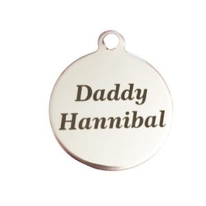 Puede incluir: Un colgante circular de color plateado con un pequeño lazo en la parte superior. El colgante tiene las palabras "Daddy Hannibal" grabadas en una fuente en negrita sin serifa. El fondo es blanco liso.