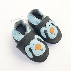May include: A pair of black leather baby shoes with a blue and orange dog design. The shoes have a soft, flexible sole and are perfect for crawling and walking.