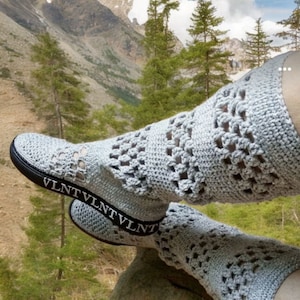 May include: Light gray crochet leg warmers and sneakers with a black sole. The leg warmers have an openwork design and extend over the sneakers. The sneakers have a black band with the text "VLNT" in white.