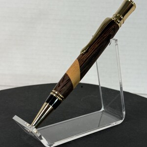 Peut inclure: Un stylo en bois artisanal avec un clip et des accents dorés. Le stylo présente un motif de grain de bois brun foncé et clair. Le stylo est présenté sur un présentoir en acrylique transparent, mettant en valeur son design et son savoir-faire.