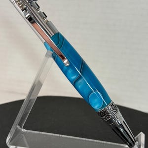 May include: A blue and silver pen with a twist mechanism and a decorative design on the end. The pen is on a clear acrylic stand.