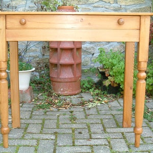 May include: A light wood table with a single drawer and turned legs. The table is in a garden setting with brick pavers and plants.