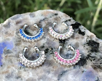 May include: Four silver nose rings with different coloured rhinestones. The rings are on a grey and white rock surface. The rhinestones are blue, clear, white, and pink.