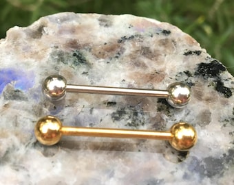 May include: Two barbell style body piercings, one silver and one gold, are shown on a light grey and white speckled surface.