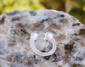 May include: A clear plastic horseshoe shaped ring with rounded ends. The ring is on a grey and white speckled surface.