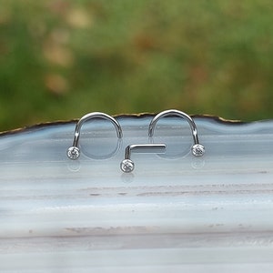 May include: Three silver nose rings with clear gemstones. Two of the rings are curved and one is straight.
