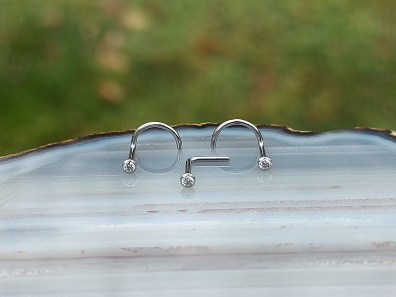 May include: Three silver nose rings with clear gemstones. Two of the rings are curved and one is straight.